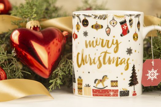 Close up of a Christmas mug with hanging empty tea label with around pine branches, red baubles and satin gold ribbon with bokeh effect