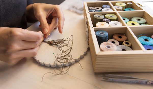 Lifestyle concept, work from home to reinvent your life: close-up of woman hands making macrame knotted jewelry with stone beads and tools on light wooden table