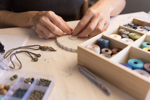 Lifestyle concept, work from home to reinvent your life: close-up of woman hands making macrame knotted jewelry with stone beads and tools on light wooden table