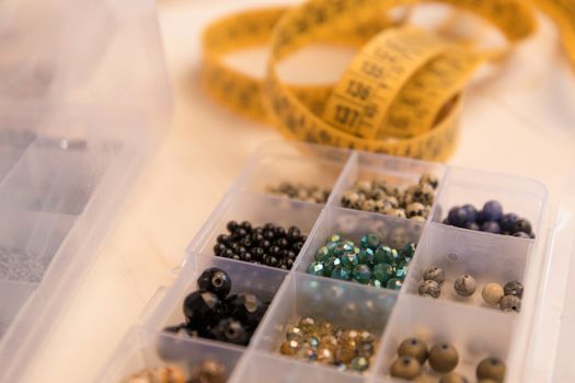 Work from home lifestyle concept: close-up detail of an organizer with various colored stone and crystal beads in foreground and a tailor meter in background bokeh effect on light wooden table