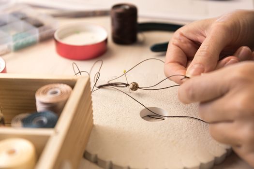 Lifestyle concept, work from home to reinvent your life: close-up of woman hands making macrame knotted jewelry with stone beads and tools on light wooden table