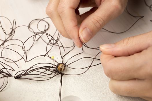 Lifestyle concept, reinvent your life and your job: close-up detail of woman hands making macrame knotted jewel with the fingers that tie the nylon thread around the diaspro natural stone