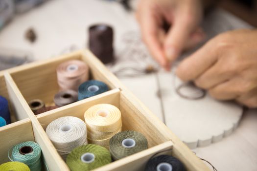 Lifestyle concept, work from home to reinvent your life: close-up detail of woman hands making macrame knotted jewel with some colored thread spools in foreground and bokeh effect