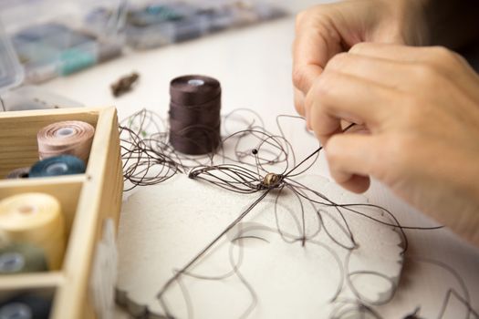 Lifestyle concept, work from home to reinvent your life: close-up detail of woman hands making macrame knotted jewel with some colored thread spools in foreground and bokeh effect