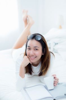 charming woman lying on bed reading a book and holding coffee cup looking into camera in bedroom.