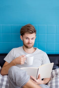 Young man reading book and drinking coffee on sofa in the living room.