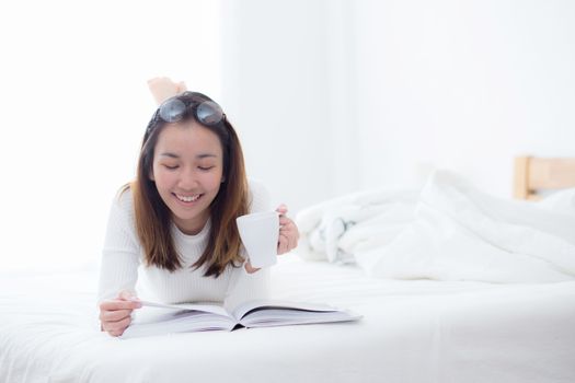 charming woman lying on bed reading a book and holding coffee cup looking into camera in bedroom.