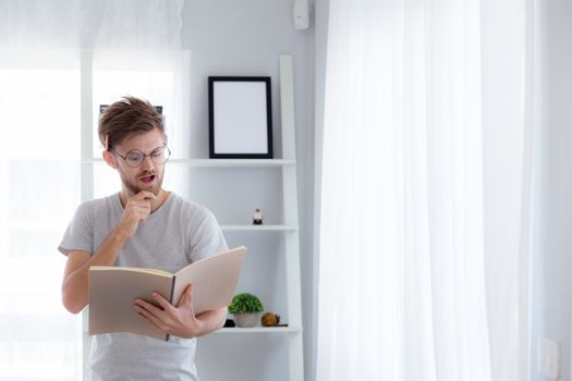 Handsome guy in eyeglasses is reading book preparing exam and thinking with standing at the living room - education concept.