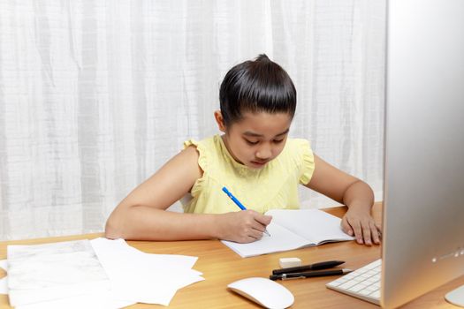 Asian little girl is studying online via the internet  sitting and writing in living room at home. Asia children writeing with pencil on notebook. online learning at home or learn frome home concept. 