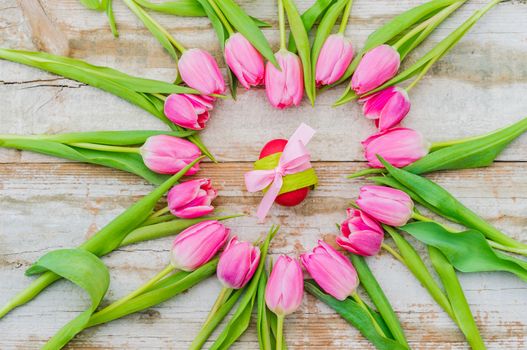 Easter decoration with egg ornated with pink ribbon bow, framed with pastel pink tulip flowers on rustic  wood