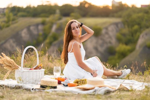 Gorgeous young brunette girl in a white sundress having a picnic in a picturesque place. Romantic picnic.