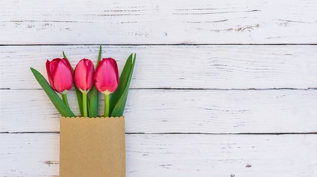Fresh red tulips in paper bag on white wooden background with copy space