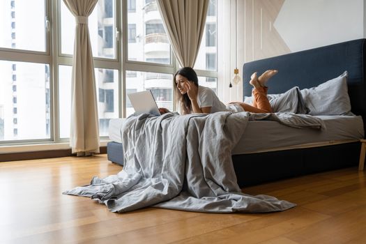 A beautiful young brunette girl is working on the laptop while lying on a bed by a panoramic window with a beautiful view from a high floor. Stylish modern interior. A cozy workplace.