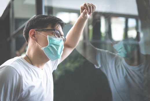 Sad young Asian man wearing hand made protective face mask looking out the window being quarantined at home. Quarantine concept during the coronavirus pandemic.