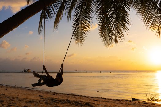 The guy enjoys the sunset riding on a swing on the ptropical beach. Silhouettes of a guy on a swing hanging on a palm tree, watching the sunset in the water