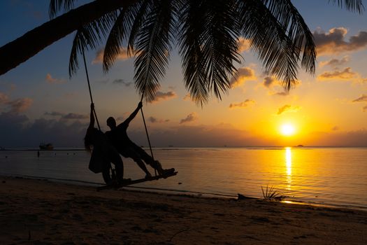 Silhouetted couple in love walks on the beach during sunset. Riding on a swing tied to a palm tree and watching the sun go down into the ocean.