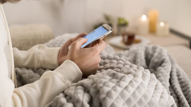 Relaxation concept: woman sitting on the sofa with a blanket on her legs using her smartphone while sliding her fingers across the screen