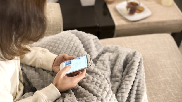 Relaxation concept: woman sitting on the sofa with a blanket on her legs using her smartphone while sliding her fingers across the screen