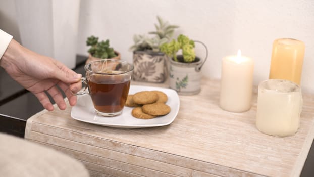 Relaxation concept: woman's hand takes a cup of tea from a plate with biscuits on a small table with candles little plants in bokeh effect
