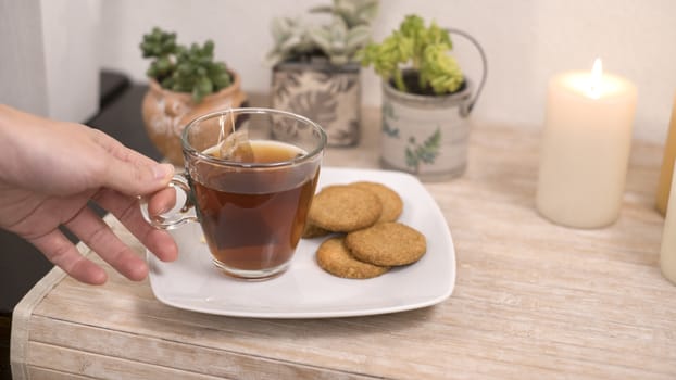 Relaxation concept: woman's hand takes a cup of tea from a plate with biscuits on a small table with candles little plants in bokeh effect