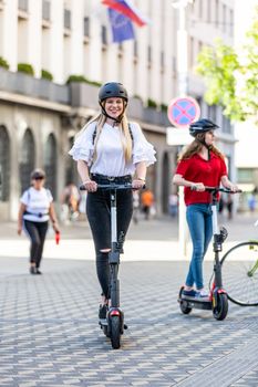 Trendy fashinable teenager girls riding public rental electric scooters in urban city environment. New eco-friendly modern public city transport in Ljubljana, Slovenia.