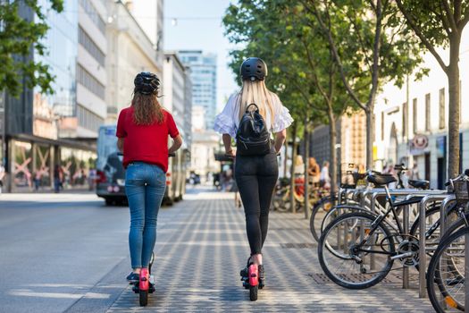 Rear view of trendy fashinable teenager girls riding public rental electric scooters in urban city environment. New eco-friendly modern public city transport in Ljubljana, Slovenia.