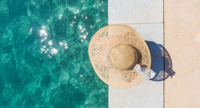 Graphic image of top down view of woman wearing big summer sun hat relaxing on pier by clear turquoise sea.