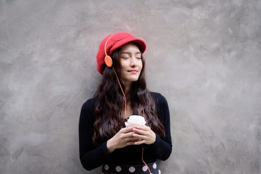 Asian portrait woman with red hat and black clothes has holding a cup of coffee with happiness and the grey stone wallpaper background.