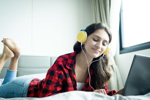 Beautiful woman working and listening to music on a laptop with yellow headphone and lying down on the bed at a condominium in the morning.