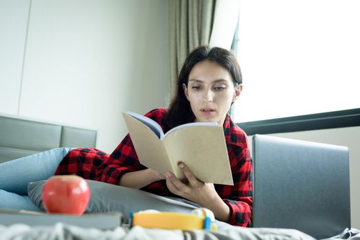 Beautiful woman working and reading a book on a laptop and lying down on the bed at a condominium in the morning.