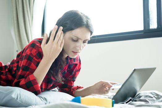 A beautiful woman has lain down on the bed at a condominium and shopping online on a laptop by the credit card with unhappy and worried in the morning.