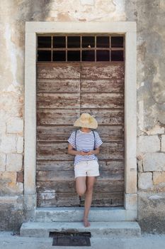 Beautiful young female tourist woman standing in front of vinatage wooden door and textured stone wall at old Mediterranean town, smiling, holding, using smart phone to network on vacationes.
