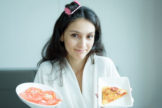 A beautiful woman wearing a towel and a white bathrobe has to eat a pizza and sliced tomato with happy and relaxing on the bed at a condominium in the morning.