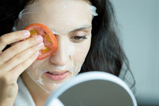 A beautiful woman wearing a towel and a white bathrobe has to look to a mirror for a mask on face-covering eyes with sliced tomatoes with happy and relaxing on the bed at a condominium in the morning.