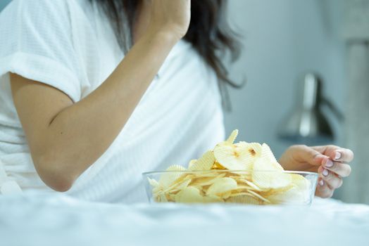 A beautiful woman eating snack potato and playing laptop with lying down on the bed and happiness at a condominium in the morning.