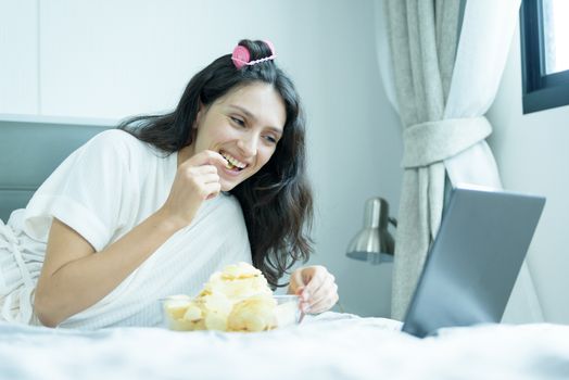 A beautiful woman eating snack potato and playing laptop with lying down on the bed and happiness at a condominium in the morning.