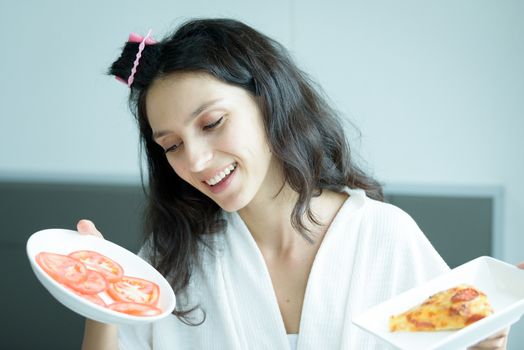 A beautiful woman wearing a towel and a white bathrobe has to eat a pizza and sliced tomato with happy and relaxing on the bed at a condominium in the morning.