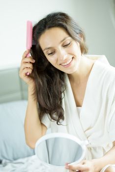 A beautiful woman wearing a towel and a white bathrobe has to look a mirror for combing hair with a pink comb and on the bed at a condominium in the morning.