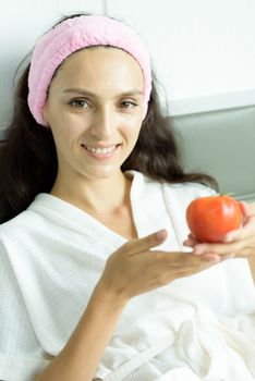 A beautiful woman wearing a towel and a white bathrobe and pink headband has to prepare to eat tomato with happy and relaxing on the bed at a condominium in the morning.