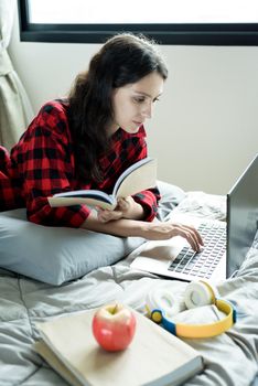 Beautiful woman working and reading a book on a laptop and lying down on the bed at a condominium in the morning.