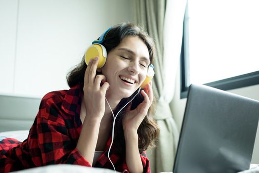 Beautiful woman working and listening to music on a laptop with yellow headphone and lying down on the bed at a condominium in the morning.