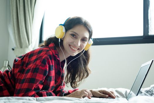 Beautiful woman working and listening to music on a laptop with yellow headphone and lying down on the bed at a condominium in the morning.