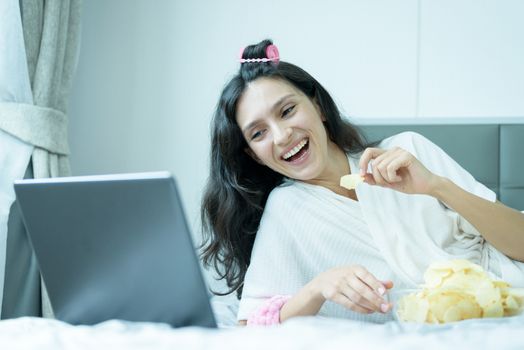 A beautiful woman eating snack potato and playing laptop with lying down on the bed and happiness at a condominium in the morning.