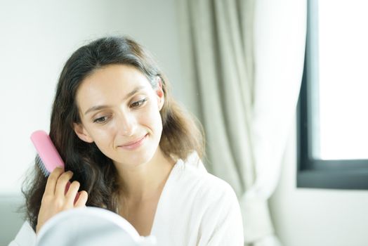 A beautiful woman wearing a towel and a white bathrobe has to look a mirror for combing hair with a pink comb and on the bed at a condominium in the morning.