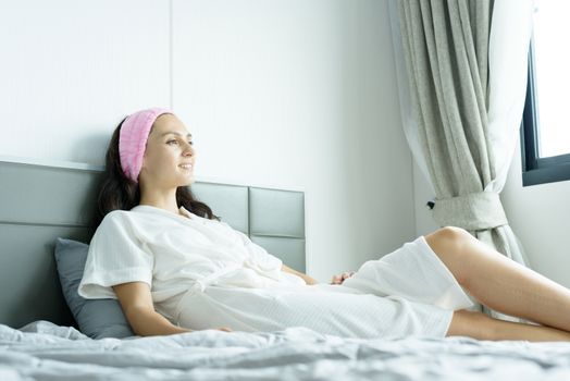 A beautiful woman wearing a towel and a white bathrobe and pink headband with happy and relaxing on the bed at a condominium in the morning.