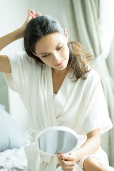 A beautiful woman wearing a towel and a white bathrobe has to look a mirror for combing hair with a pink comb and on the bed at a condominium in the morning.