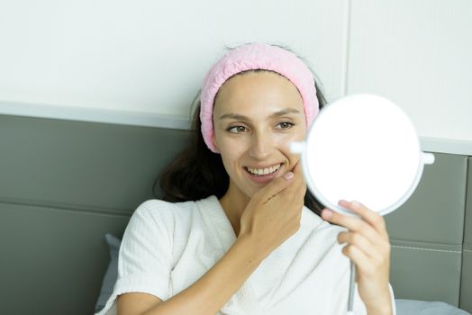 A beautiful woman wearing a towel and a white bathrobe and pink headband has to look a mirror with happy and relaxing on the bed at a condominium in the morning.