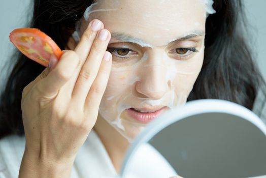 A beautiful woman wearing a towel and a white bathrobe has to look to a mirror for a mask on face-covering eyes with sliced tomatoes with happy and relaxing on the bed at a condominium in the morning.