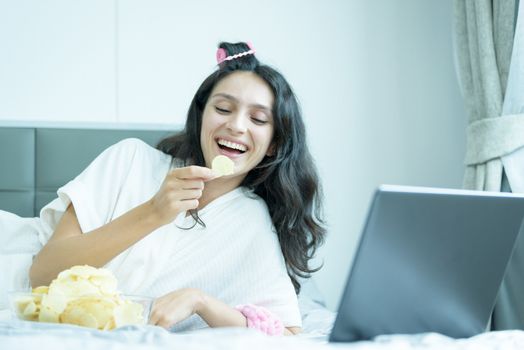 A beautiful woman eating snack potato and playing laptop with lying down on the bed and happiness at a condominium in the morning.