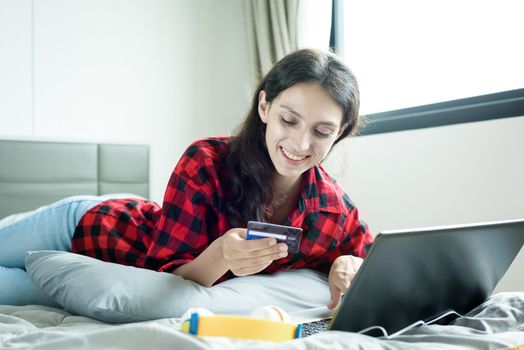 Beautiful woman shopping online on a laptop with the credit card and lying down on the bed at a condominium in the morning.